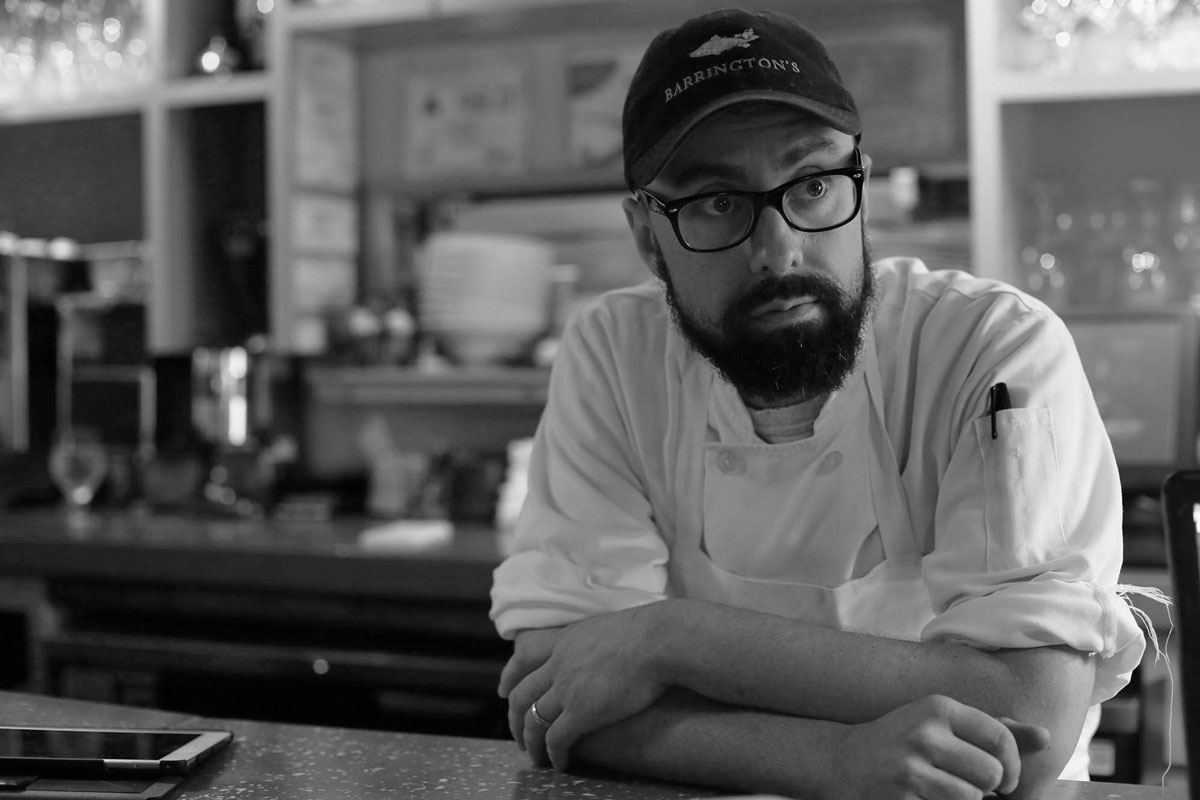 chef leaning over a counter with beard, glasses, and hat